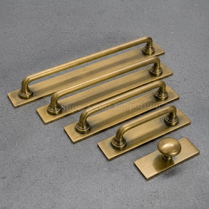 May include: A collection of five antique brass hardware pieces, including four rectangular drawer pulls of varying lengths and one single round knob, each mounted on a matching rectangular backplate with a brushed metallic finish.