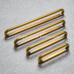 May include: Four elongated, arch-shaped cabinet pulls in a brushed brass finish, arranged diagonally from longest to shortest. Each handle features a smooth, continuous metal loop design with rounded edges and a warm, metallic gold-toned surface.