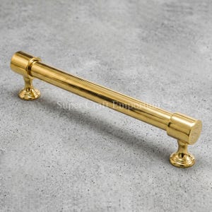 May include: A polished gold-tone cabinet pull with a cylindrical bar and decorative end pieces. The hardware is set against a gray background. The text "Super Craft Emporium" is visible on the surface.