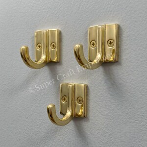 May include: Three polished brass wall hooks arranged in a triangular formation. Each hook features a rectangular base plate with two visible Phillips head screws and a curved hook extending outward. The metal has a reflective, smooth finish.