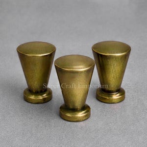 May include: Three brass cabinet knobs with a conical shape and a rounded top. The knobs have a vintage, antique appearance and are set against a grey background. The base of each knob is circular.