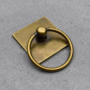 May include: Antique brass cabinet pull with a square backplate and a circular ring handle. The hardware has a vintage aesthetic, suitable for furniture restoration or adding a classic touch to new cabinetry. The pull is made of metal and has a warm, aged finish.