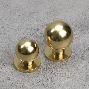 Peut inclure: Deux boutons de meuble dorés et sphériques. Chaque bouton a une base ronde et une surface lisse et réfléchissante. Les boutons sont en métal et sont probablement destinés aux meubles ou à la décoration. Le texte "Supercraft Emporium" est visible.