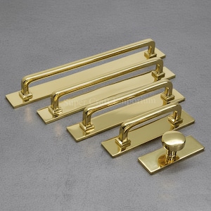May include: A selection of gold-coloured cabinet hardware, featuring rectangular bar pulls and a round knob. The hardware is displayed on a grey surface, highlighting the different sizes and styles for home improvement.