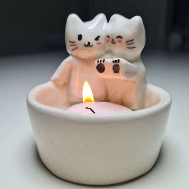 Pu&ograve; includere: Portacandele in ceramica bianca con due gatti in ceramica bianca che si tengono per mano. I gatti hanno occhi neri e baffi. Una piccola candela da t&egrave; &egrave; accesa all'interno del portacandele.