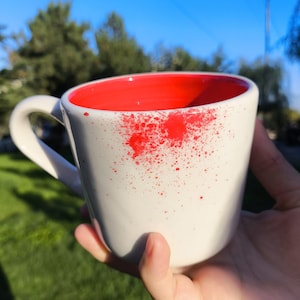 May include: A white ceramic mug with a red interior and red splatter design on the exterior. The mug has a curved handle and is held against a background of green grass and a blue sky.