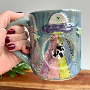 Puede incluir: Taza de cerámica turquesa con un diseño en relieve de un OVNI, un extraterrestre y una vaca abducida. La taza tiene un asa y está decorada con estrellas y rayas de colores. El dije de vaca cuelga del OVNI.