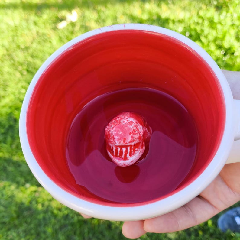 May include: White ceramic mug with a red interior. A red, textured dinosaur head is centered inside the mug. The mug is filled with a red liquid. The mug is held by a person.
