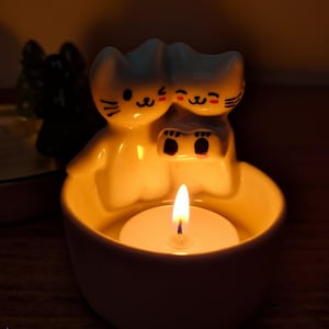 Pu&ograve; includere: Portacandele in ceramica bianca a forma di due gatti con una candela tea light all'interno. I gatti si guardano e hanno occhi neri e baffi.