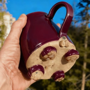 May include: A handmade ceramic mug in a deep burgundy glaze, held in a hand. The base features four rounded feet and a decorative design. The mug has a large handle and a unique, artistic design.