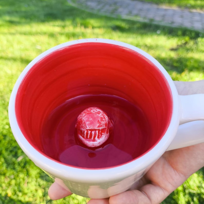 May include: White ceramic mug with a red interior and a sculpted, red, creature-like design inside. The mug has a white handle and a white exterior. The mug is held in a hand against a background of green grass.