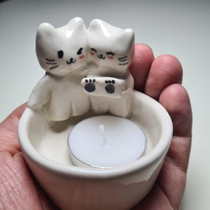 Pu&ograve; includere: Portacandele in ceramica bianca con due gatti carini che tengono una candela da t&egrave;. I gatti hanno occhi neri e baffi, e il portacandele ha una glassa bianca.