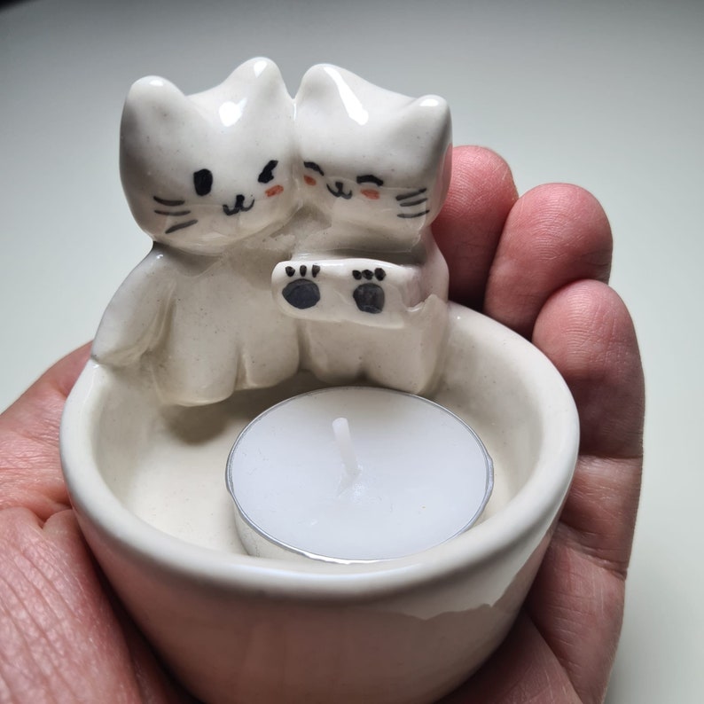 Pu&ograve; includere: Portacandele in ceramica bianca con due gatti carini che tengono una candela da t&egrave;. I gatti hanno occhi neri e baffi, e il portacandele ha una glassa bianca.
