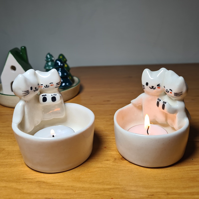 Pu&ograve; includere: Due portacandele in ceramica bianca a forma di gatto. I gatti tengono una candela a t&egrave; in ogni portacandele. I portacandele sono su una superficie di legno.