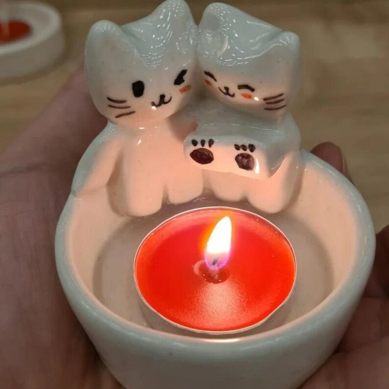 Pu&ograve; includere: Portacandele in ceramica bianca con due gatti carini che si tengono le zampe. Il portacandele ha una piccola depressione rotonda al centro per contenere una candela da t&egrave;. Una candela da t&egrave; rossa &egrave; accesa all'interno del portacandele.