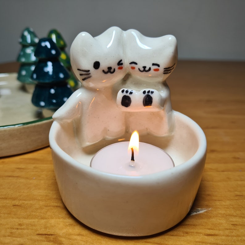 Pu&ograve; includere: Portacandele in ceramica bianca a forma di due gatti che si tengono le zampe, con una piccola candela bianca all'interno. Il portacandele &egrave; su una superficie di legno.
