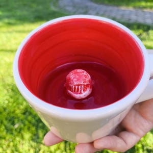 May include: White ceramic mug with a red interior. A small, red, shark-shaped figure is centered inside the mug. The mug is held against a blurred green background.