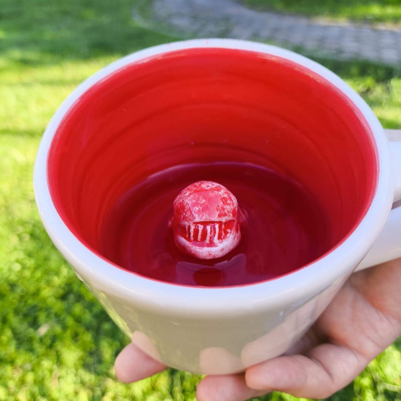 May include: White ceramic mug with a red interior. A small, red, shark-shaped figure is centered inside the mug. The mug is held against a blurred green background.