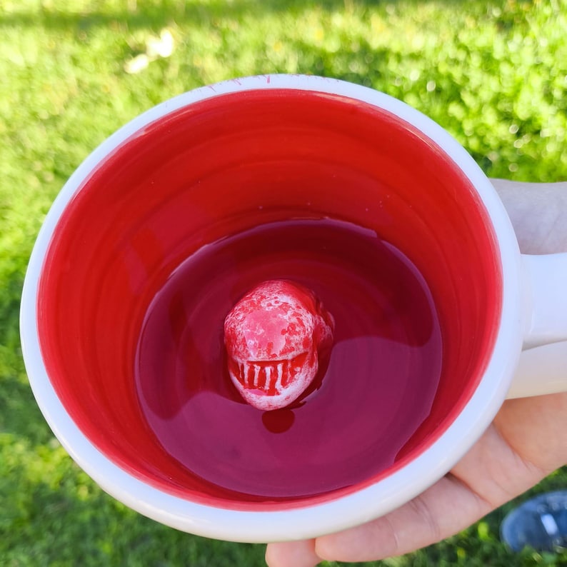May include: White ceramic mug with a red interior. A red, textured, three-dimensional alien head is inside the mug. The mug is filled with a red liquid. The mug is held by a person.