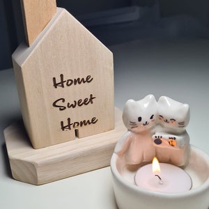 Pu&ograve; includere: Un portacandele in legno a forma di casa con la scritta "Home Sweet Home" incisa sul davanti. Il portacandele ha una statuetta di gatto in ceramica con una candela a t&eacute; all'interno.