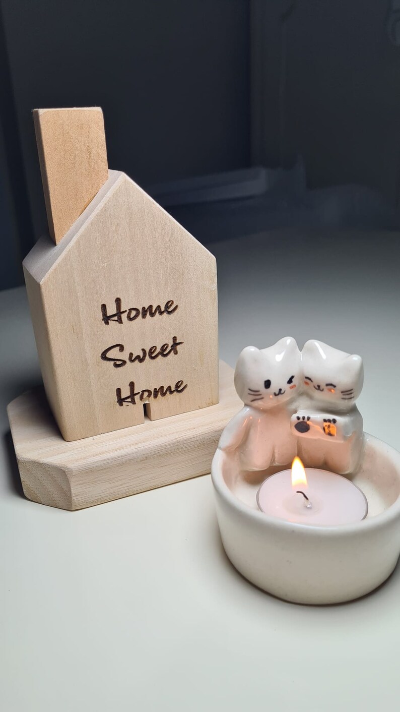 Pu&ograve; includere: Un portacandele in legno a forma di casa con la scritta "Home Sweet Home" incisa sul davanti. Il portacandele ha una statuetta di gatto in ceramica con una candela a t&eacute; all'interno.