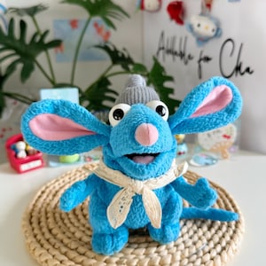 May include: A blue plush mouse toy with large ears, pink inner ears, and a grey knitted cap. The mouse has a cream-coloured scarf and sits on a round, woven placemat. The background includes greenery and decorative items.