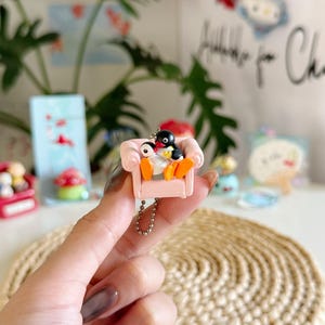 Puede incluir: Un llavero en miniatura con dos figuras de pingüinos sentados en un sofá rosa. Los pingüinos son negros, blancos y amarillos. El sofá tiene cojines naranjas. El llavero está sujeto a una cadena plateada. El fondo está borroso, con otros pequeños objetos decorativos.