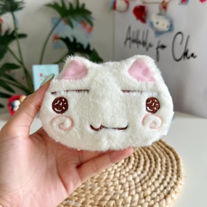 Pode incluir: Uma bolsa de pelúcia branca em formato de rosto de gato com pelo macio. O rosto apresenta detalhes bordados, incluindo olhos fechados, orelhas internas cor-de-rosa, pupilas circulares castanhas e bochechas rosadas em espiral com boca.