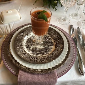 May include: A table setting with a white tablecloth, a plate with a brown and white floral pattern, a pink and white patterned charger plate, and a glass of tomato soup with a basil garnish.