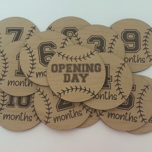 May include: A collection of wooden baseball-themed milestone discs. Each disc is round with a baseball stitch design and a number or text indicating a month or special occasion. The text "Opening Day" is featured on one disc.