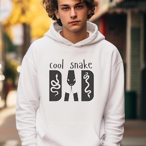 Könnte beinhalten: Weißer Kapuzenpullover mit dem Aufdruck "cool snake dad" auf der Vorderseite. Der Text ist schwarz und zeigt ein Schlangendesign.