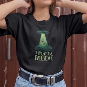 May include: Black t-shirt with a graphic of a UFO beaming up a reptile over a forest scene. The text "I WANT TO BELIEVE" is printed in green below the image. The shirt is made of a soft material.
