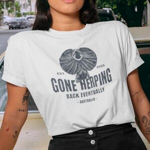 May include: White t-shirt featuring a frilled-neck lizard graphic and the text "GONE HERPING BACK EVENTUALLY - AUSTRALIA - EST. 2025". The design is in grey and black.