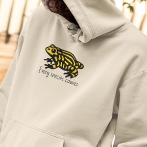 Może przedstawiać: Kremowa bluza z kapturem z żółto-czarną ilustracją żaby. Pod żabą wydrukowano napis "Every species counts". Bluza ma przednią kieszeń i kaptur ze sznurkiem.