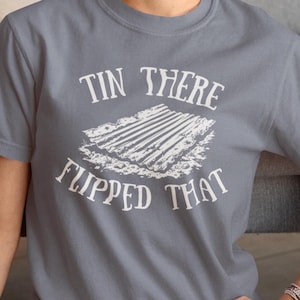 Può includere: T-shirt grigia con la scritta bianca "Tin There Flipped That" sopra una grafica bianca di lamiere ondulate. La maglietta ha maniche corte e girocollo.