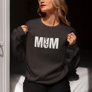 Könnte beinhalten: Ein dunkelgrauer Pullover mit dem Wort "MUM" in Weiß gedruckt, wobei das "U" mit einem stilisierten Kringel verziert ist.