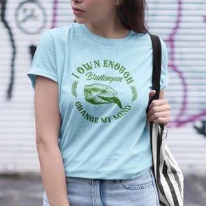 May include: Light blue t-shirt with the text "I OWN ENOUGH Bluetongues CHANGE MY MIND" in a circular design. The design includes a green illustration of a blue-tongued skink. The shirt is made of a soft material.