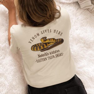 May include: A cream-coloured t-shirt with a detailed illustration of an Eastern Tiger Snake. The text reads "VENOM LIVES HERE" and the scientific name "Notechis scutatus". The snake is in yellow, brown, and black.