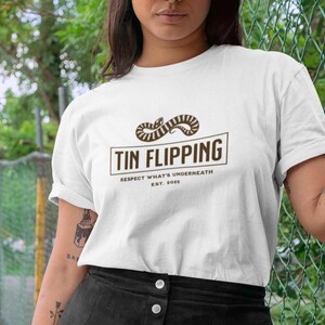 May include: White t-shirt with the text "TIN FLIPPING" in a brown rectangular box, above the text "RESPECT WHAT'S UNDERNEATH EST. 2025". The shirt features a brown snake design above the text. The shirt is made of a soft material.
