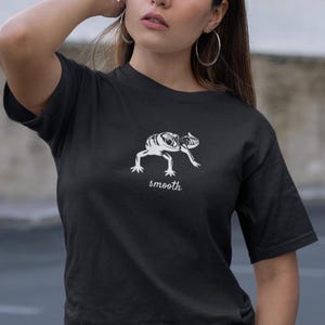 Op de afbeelding: Zwart T-shirt met een witte illustratie van een hagedisachtig wezen. Het woord "smooth" is in cursieve letters onder de afbeelding geschreven. Het shirt heeft korte mouwen en een ronde hals.
