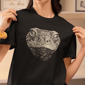 May include: Black t-shirt featuring a detailed, grayscale illustration of a reptile's face. The image showcases the textured skin and expressive eyes of the creature. The design is centered on the shirt, creating a striking visual.