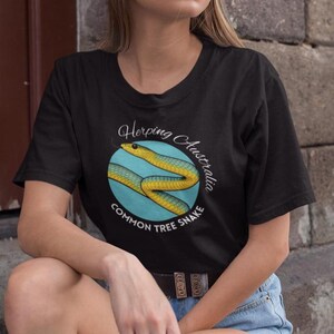 May include: Black t-shirt with a graphic of a yellow and blue Common Tree Snake inside a blue circle, with the text "Herping Australia" and "Common Tree Snake". The shirt has a crew neck and short sleeves.