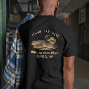 May include: Black t-shirt with a gold illustration of an Inland Taipan snake, with the text "VENOM LIVES HERE" above and "Oxyuranus microlepidotus -INLAND TAIPAN-" below. The design features a coiled snake.