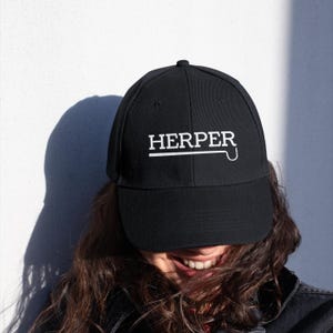 Puede incluir: Gorra de béisbol negra con la palabra "HERPER" en letras blancas. La gorra tiene una visera curva y un ajuste estructurado. El texto está centrado en la parte delantera, con una línea blanca y una "J" estilizada debajo.
