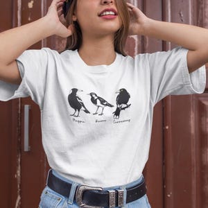 May include: White t-shirt with three bird illustrations: a magpie, a pewee, and a currawong. Each bird is in black and white, with its name below. The shirt has short sleeves and is made of a soft material.