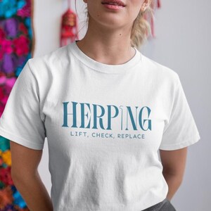 Op de afbeelding: Wit t-shirt met het woord "HERPING" in blauwgroen, samen met de tekst "LIFT, CHECK, REPLACE" eronder. Het shirt heeft korte mouwen en een ronde hals. De afbeelding toont het bovenlichaam van een persoon die het shirt draagt.