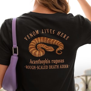 Op de afbeelding: Zwart t-shirt met een slangafbeelding en de tekst "VENOM LIVES HERE". De slang is bruin en beige, met de tekst "Acanthophis rugosus - ROUGH-SCALED DEATH ADDER -" eronder.