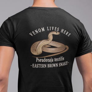 May include: Black t-shirt with a detailed illustration of an Eastern Brown Snake, featuring the text "Venom Lives Here" and the scientific name "Pseudonaja textilis". The snake is illustrated in shades of brown and tan.