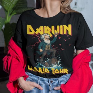 Puede incluir: Camiseta negra con un gráfico de Charles Darwin en pose de rock and roll, con la palabra "DARWIN" en letras amarillas y rojas encima. Las palabras "WORLD TOUR" están debajo.