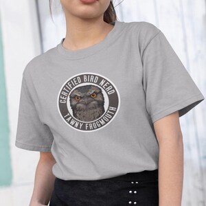 Puede incluir: Una camiseta gris claro con un gráfico circular. El gráfico dice "CERTIFIED BIRD NERD TAWNY FROGMOUTH" y presenta una ilustración detallada de un pájaro lechuzón con ojos anaranjados.